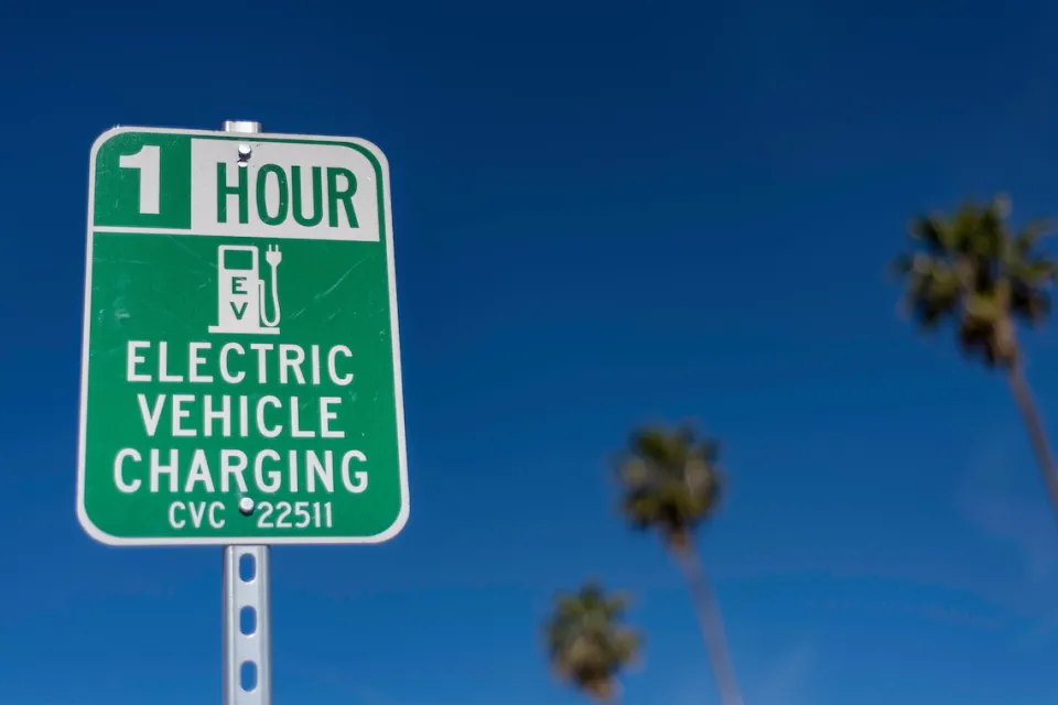 Close-up of green and white sign for 1 hour EV charging station outdoors with tall palm trees in background against blue sky.